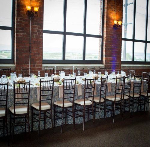 Long rectangular dining table set for event; brown Chiavari chairs, neutral tablecloth, brick wall, large windows.