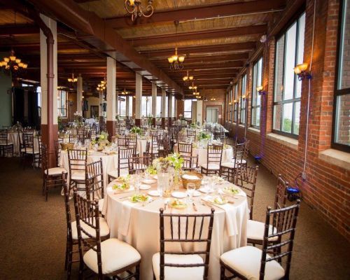 Wedding reception in a brick-walled room with round tables set for guests, sunlight through large windows.