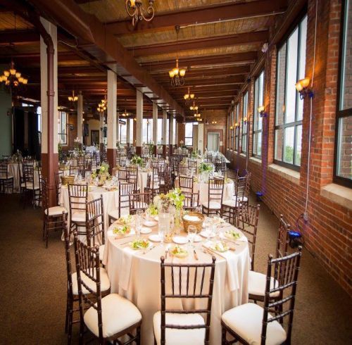 Wedding reception in a brick-walled room with round tables set for guests, sunlight through windows.