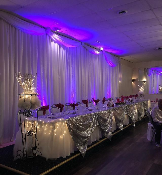 Banquet table set for an event; draped with white and silver fabric, purple uplighting.