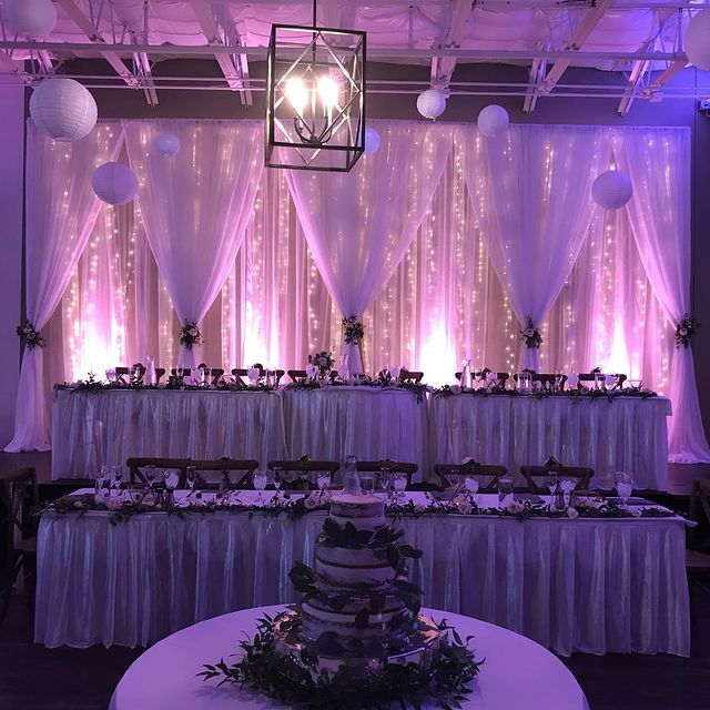 Wedding reception with draped backdrop, tables, cake, and floral arrangements, illuminated with pink lighting.