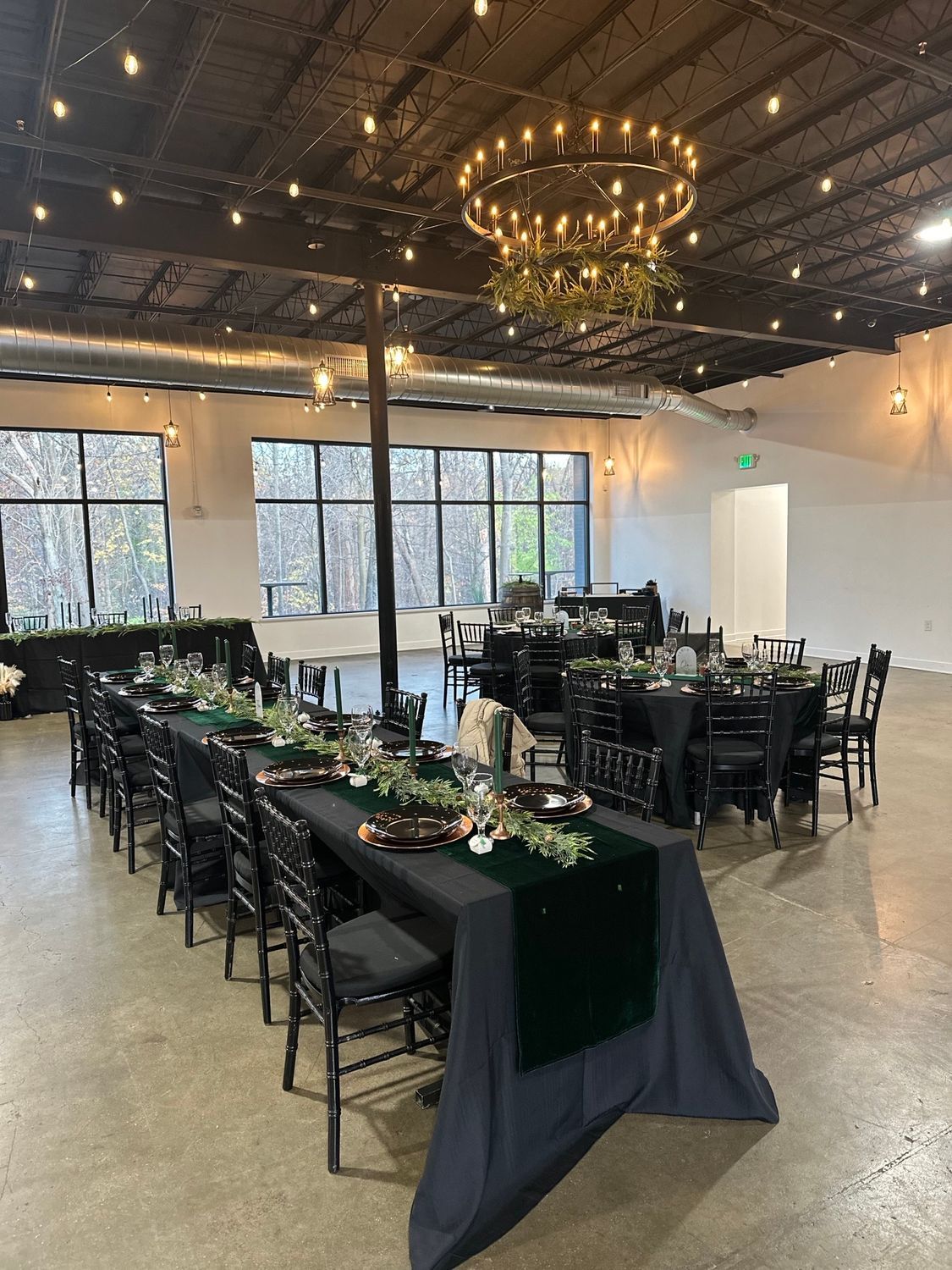 Event hall with long and round tables set for a dinner, black chairs and linens, greenery centerpieces.