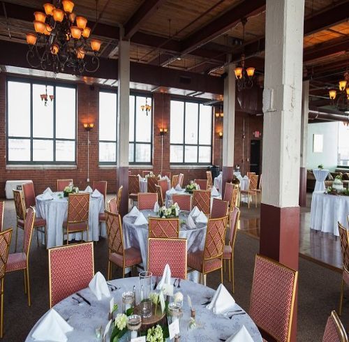 Reception hall with round tables, chairs, and ornate chandeliers. Windows and brick walls.