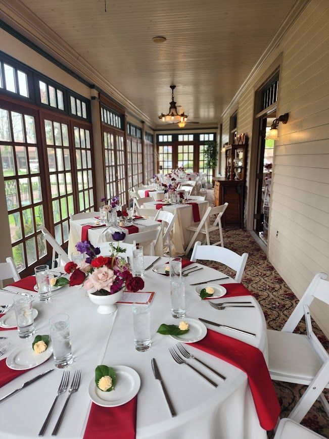 Long, light-filled dining room set for a celebration. Round tables with red napkins and floral centerpieces.