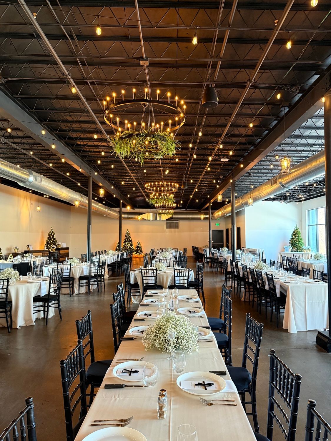 Wedding reception in a large room with long tables set for guests; overhead string lights and chandeliers.