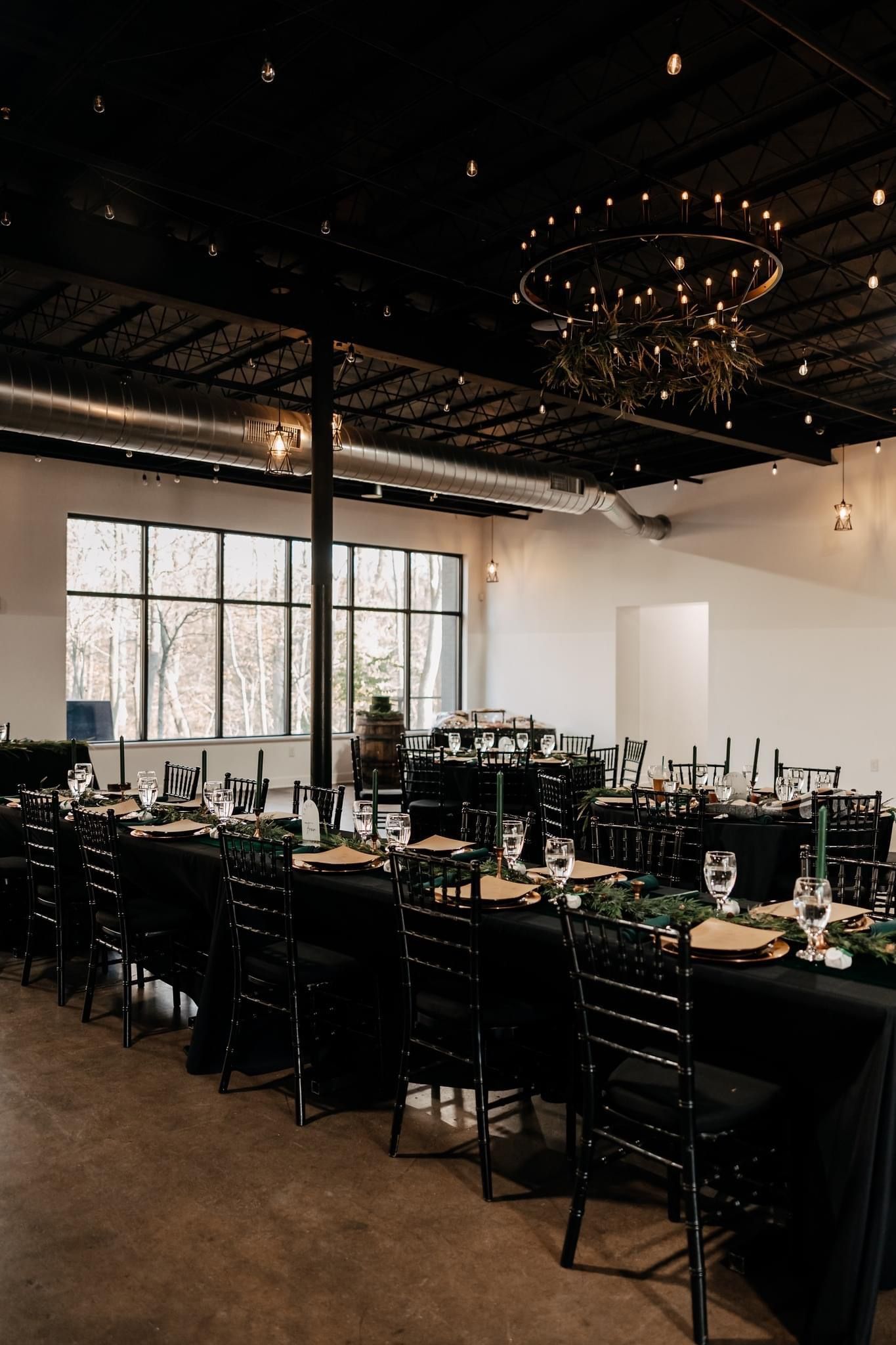 Black-decorated event hall with tables set for a wedding. Greenery and gold accents complement the dark decor.