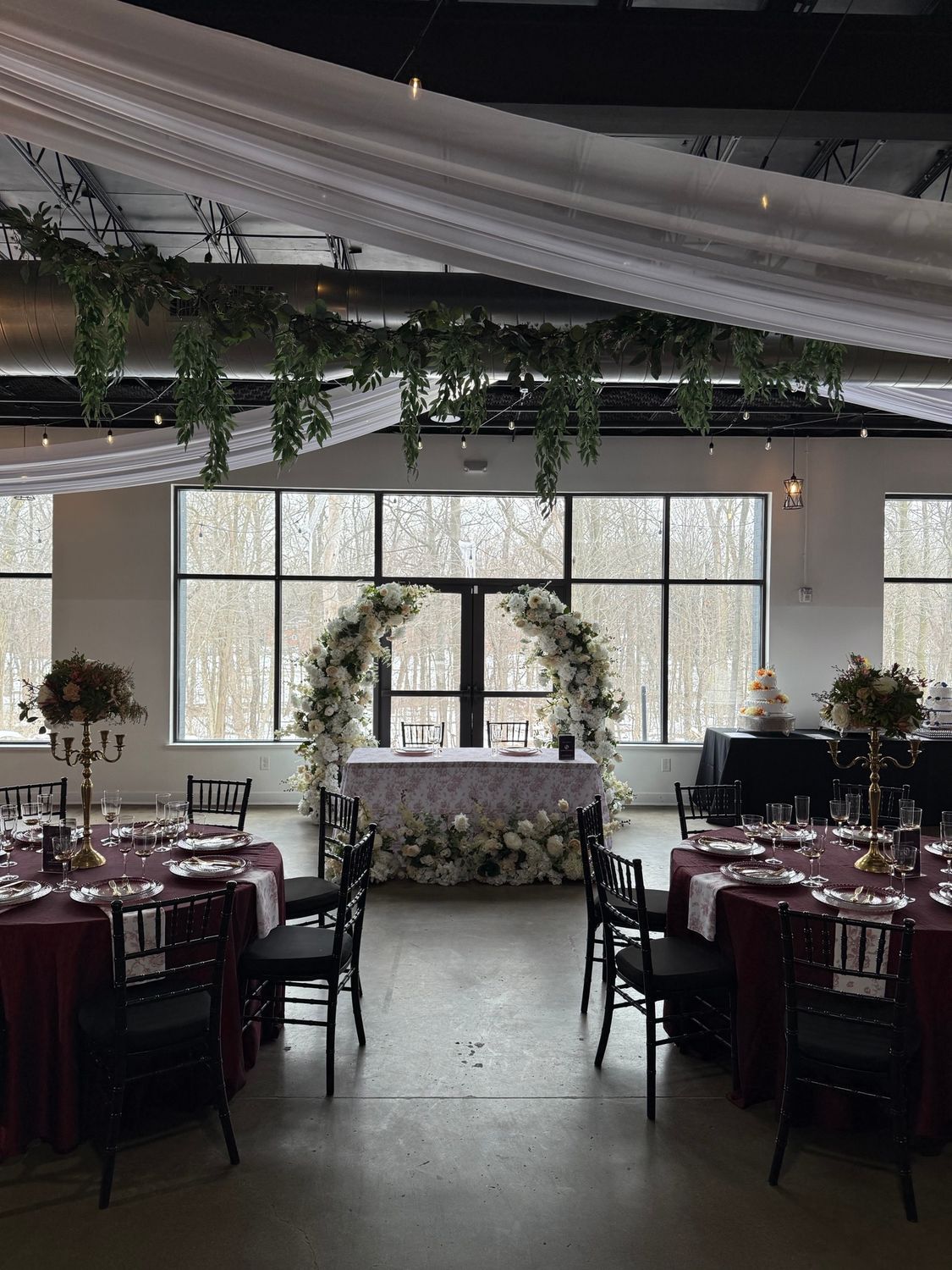 Wedding reception with tables, floral arch, and greenery hanging from the ceiling. Burgundy tablecloths, black chairs, and large windows.