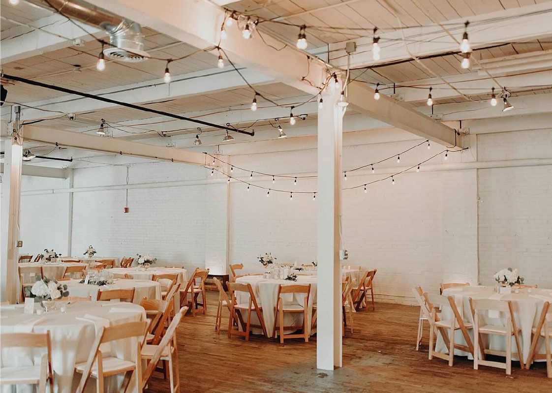 Event space with round tables, chairs, and string lights. White walls and wooden floor.