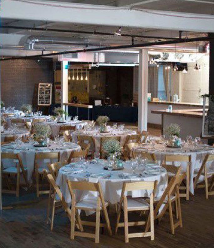 Tables set for an event inside a spacious, industrial-style room with beige chairs and white tablecloths.