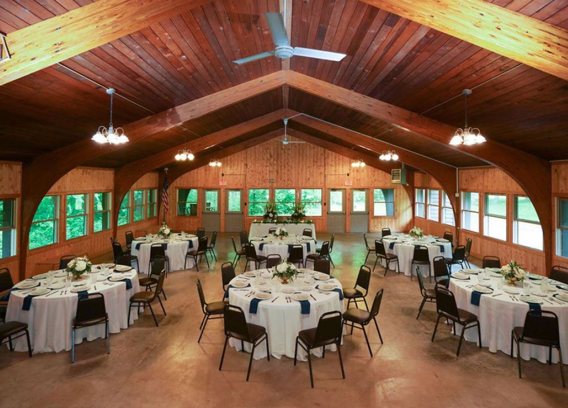 Reception hall with round tables set for a meal, under a wood-beamed ceiling. Windows look out onto greenery.