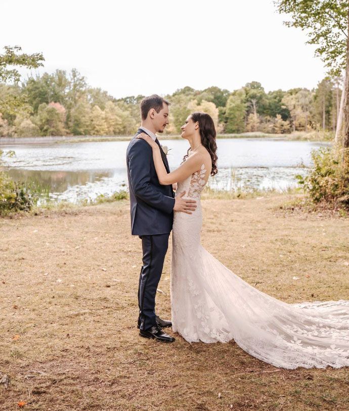 Bride and groom embrace by a lake. The bride wears a white lace dress with a long train, groom in a navy suit.