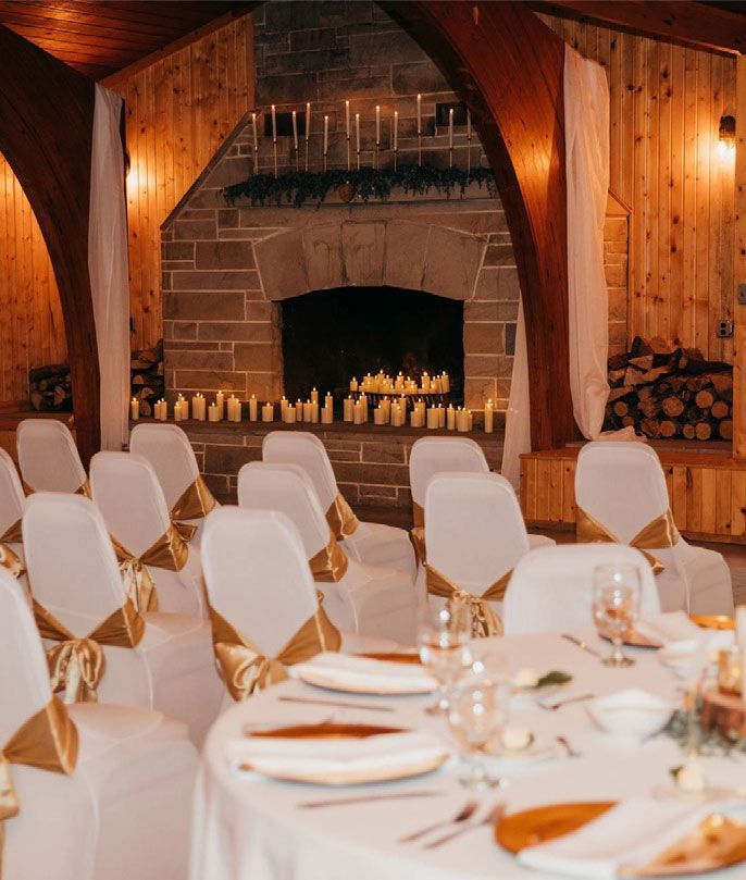 Wedding ceremony setup with white chairs, gold sashes, and lit candles near a stone fireplace.