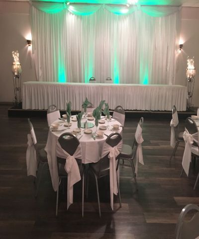 Wedding reception hall with white draped backdrop, green uplighting, and decorated tables.