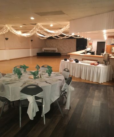 A banquet hall set for a gathering. Tables are set with white linens and green napkins. Fairy lights hang from the ceiling.