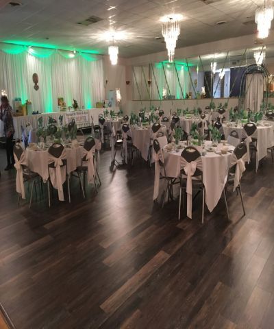 A banquet hall with round tables set for a celebration. Green and white decor with dark wood floor.