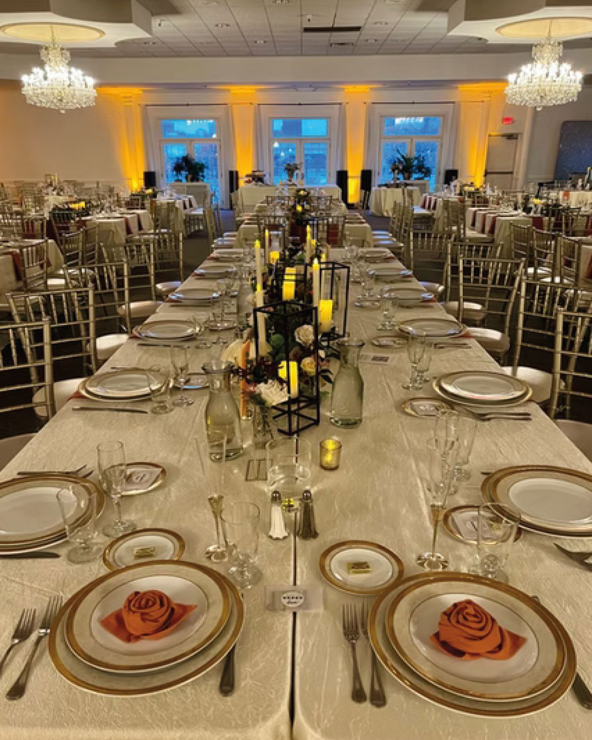 Long table set for a formal event with gold-trimmed plates, centerpieces, and warm lighting.