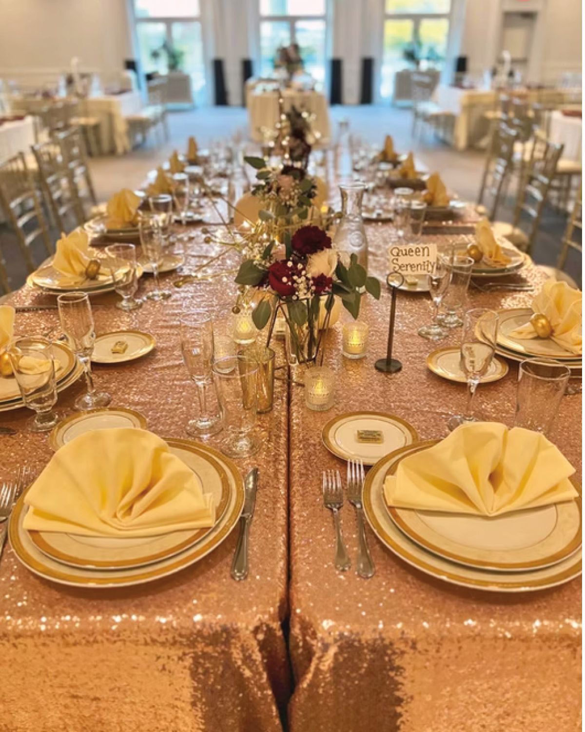 Long table set for a formal event with gold-toned sequin tablecloth, place settings, and floral centerpieces.