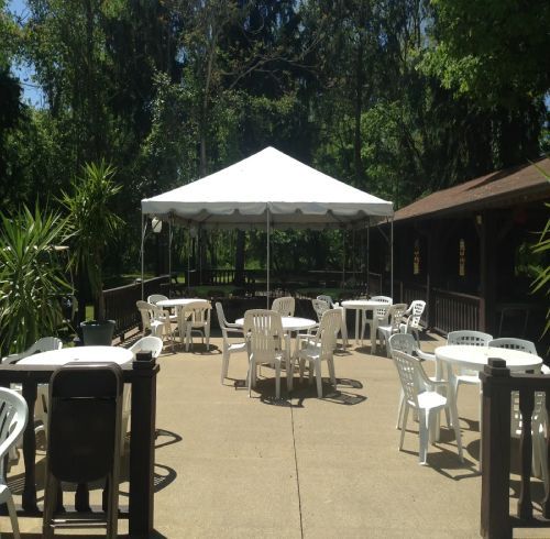 Outdoor patio with white tables and chairs under a white tent; trees in the background.