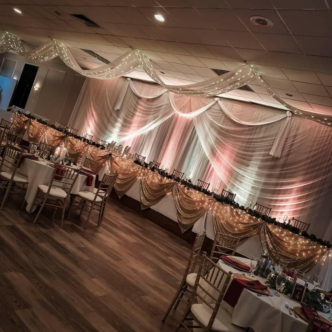 Wedding reception hall with draped white and gold fabric, fairy lights, and long banquet tables.