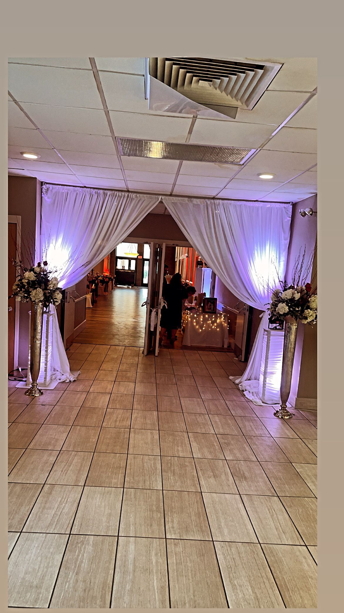A decorated hallway with white draped fabric, floral arrangements, and ambient lighting, leading to a room.