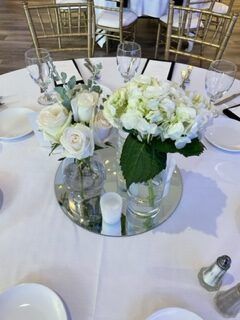 Wedding table setting with white tablecloth, floral centerpieces, glassware, and gold chairs in the background.