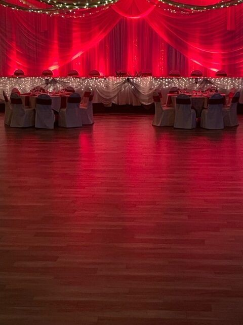 Red-lit banquet hall with decorated tables and chairs, wood floor, and draped red backdrop.