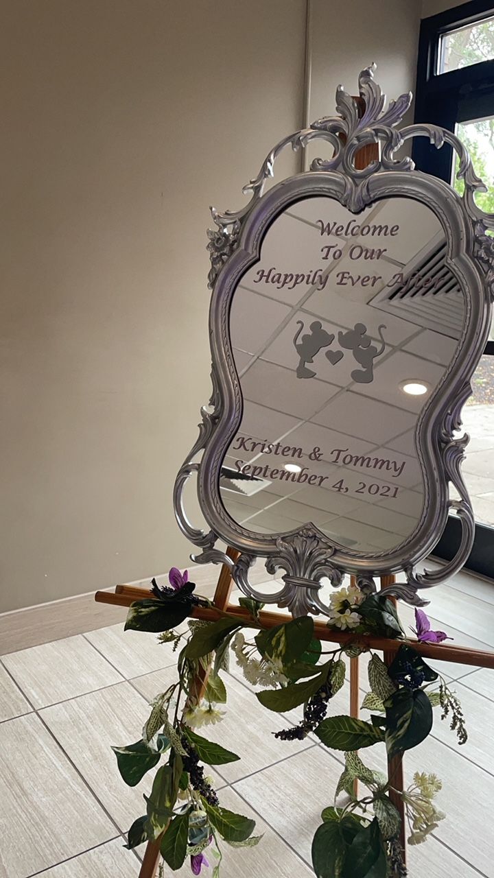 Silver framed mirror wedding welcome sign. Floral garland, text: Welcome to Our Happily Ever After & names/date.