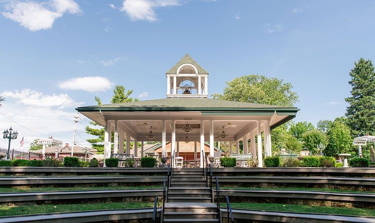 White gazebo with green roof in a park, tiered seating in foreground, blue sky.
