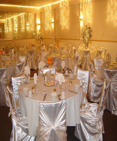 Elegant banquet hall set for a celebration. White tablecloths, chair covers, and string lights create a festive atmosphere.