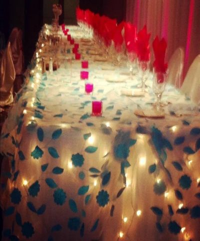 Long table set for a celebration, decorated with lights, teal and white cloth, red napkins and candles.