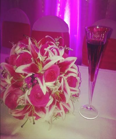 Bouquet of pink and white flowers next to a tall glass of red liquid on a white tablecloth; chairs with red sashes in the background.