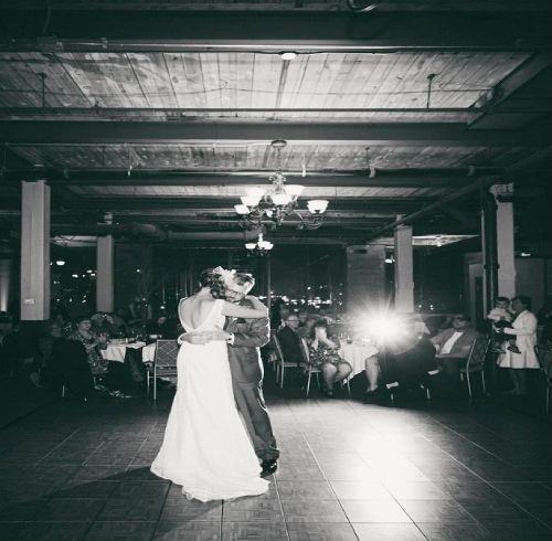 Bride and groom dance at their wedding reception; guests seated at tables under exposed beams.