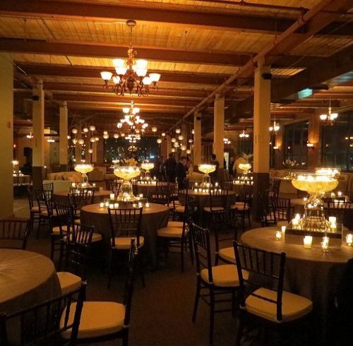 Warmly lit event space with round tables, chairs, and chandeliers. Candlelit ambiance.