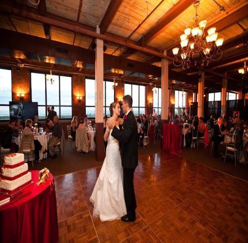 Bride and groom dance at wedding reception in an industrial-style venue, with guests and a tiered cake.