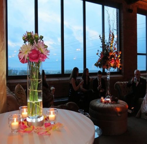 Round table with candles and flower arrangement, people seated near windows overlooking an evening view.