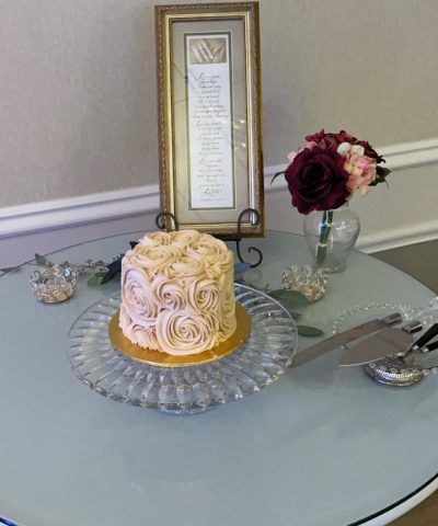 Small cake with frosting roses on a glass stand, flowers, framed poem, and cake-cutting set on a table.