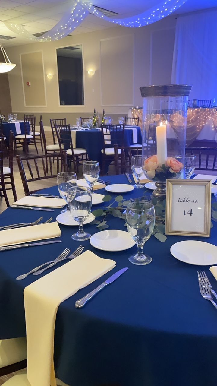 Wedding reception table set with blue tablecloth, white plates, silverware, and floral centerpiece.