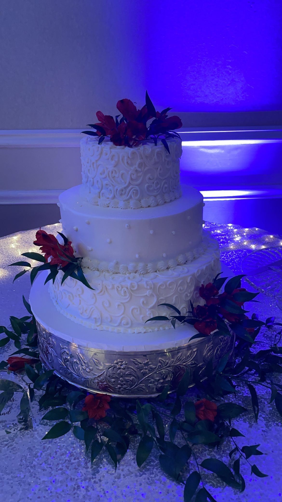 Three-tiered white cake, decorated with red flowers and greenery, on a silver platter, with blue lighting.