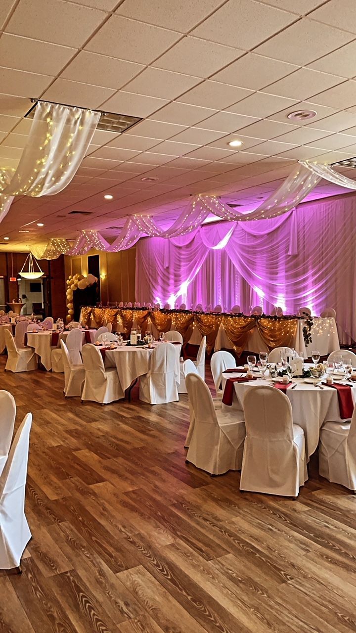 Wedding reception hall with tables set for guests, draped in white and burgundy, with soft pink lighting.