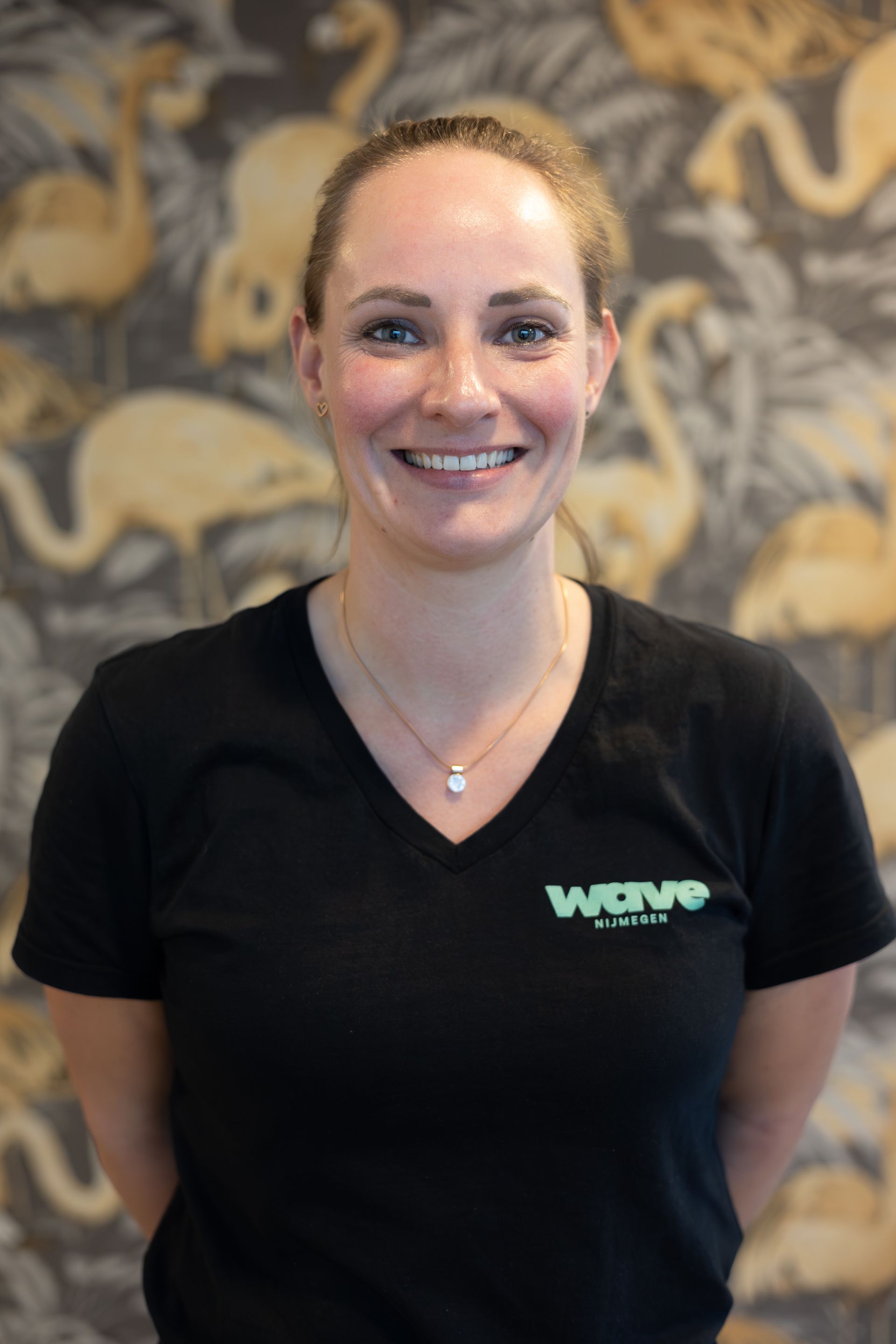 Lachende vrouw Agnes in een zwart T-shirt met V-hals en logo, staand binnen tegen een muur met patroon.