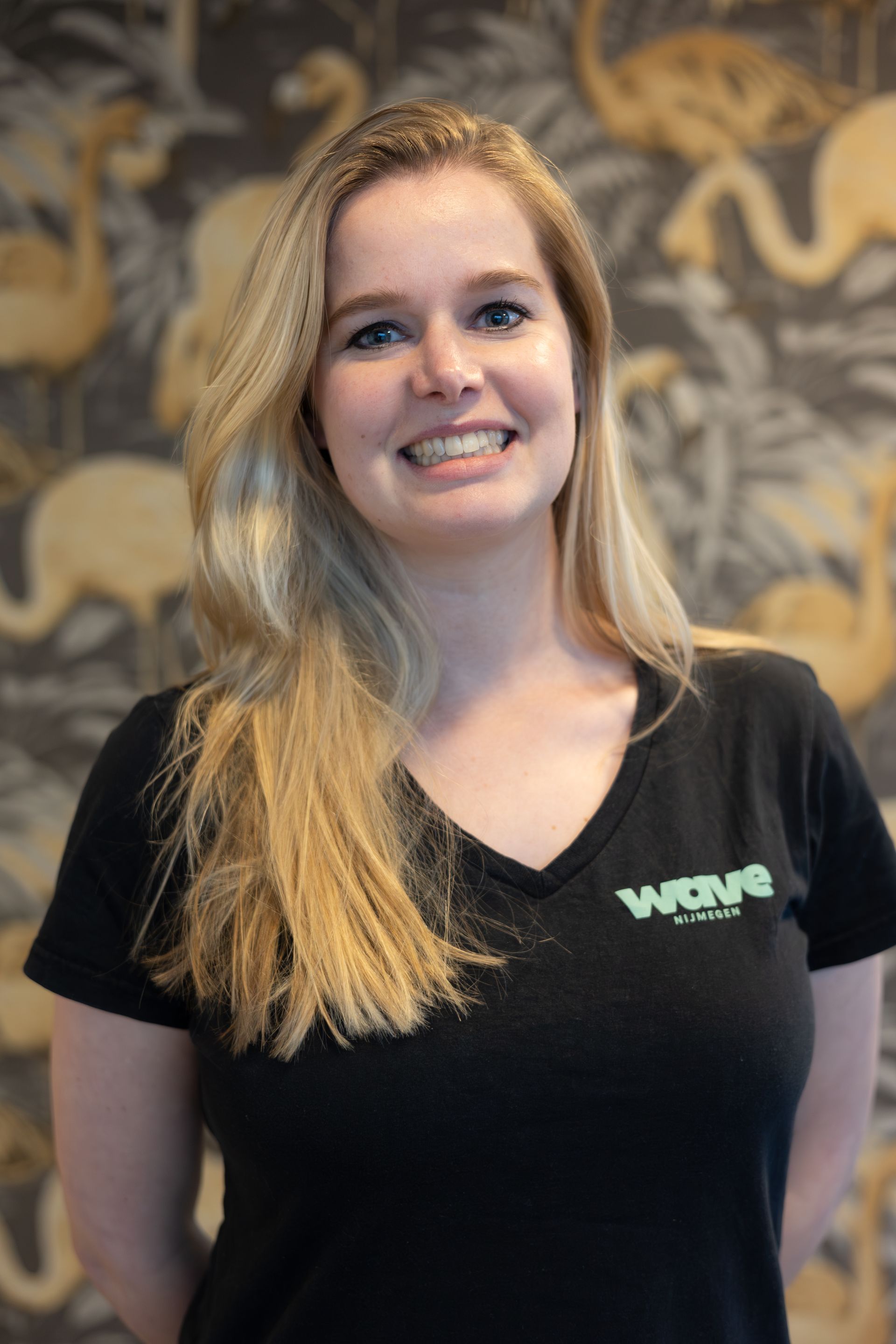 Lachende vrouw Fleur in een zwart Wave T-shirt staat voor een muur met een patroon.