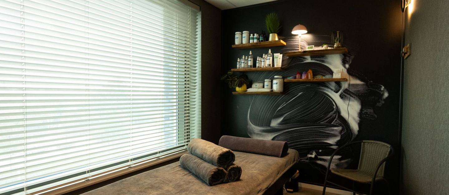 Gezellige behandelkamer met een massagetafel, opgerolde handdoeken, planken en een groot raam met jaloezieën.