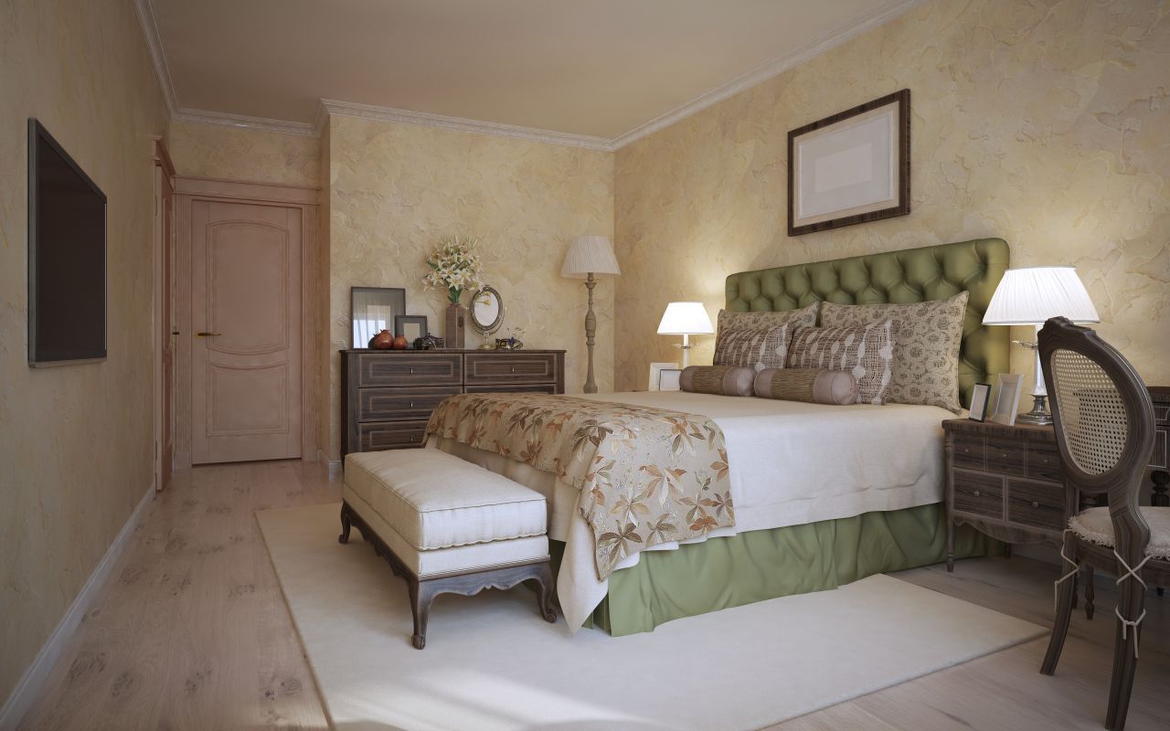 Interior of a home with arched doorways, warm yellow walls, wood floors, and potted plants. Elegant beige bedroom with green-upholstered bed, two lamps, dresser, bench, and desk chair