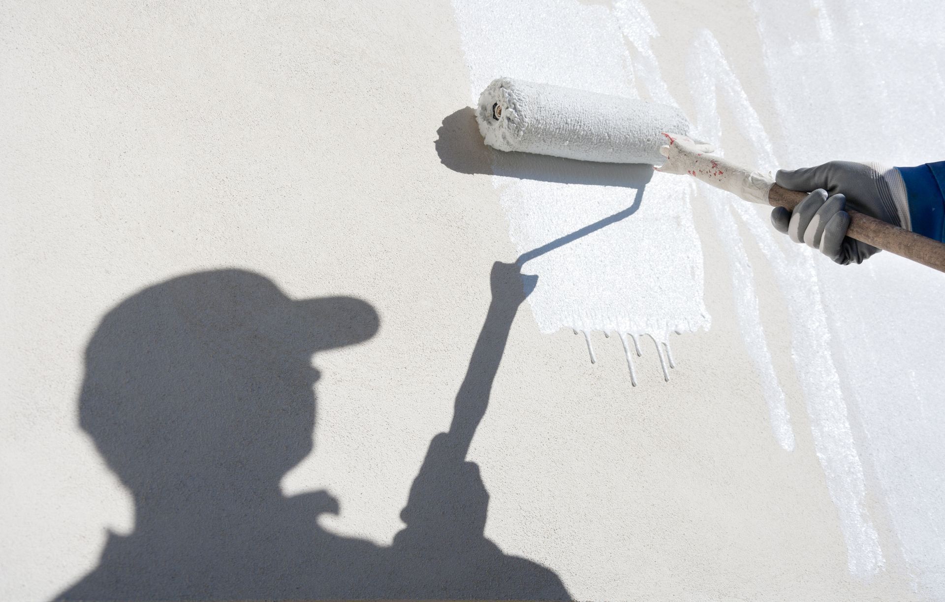 Person painting a white wall with a roller, casting a shadow in the sunlight