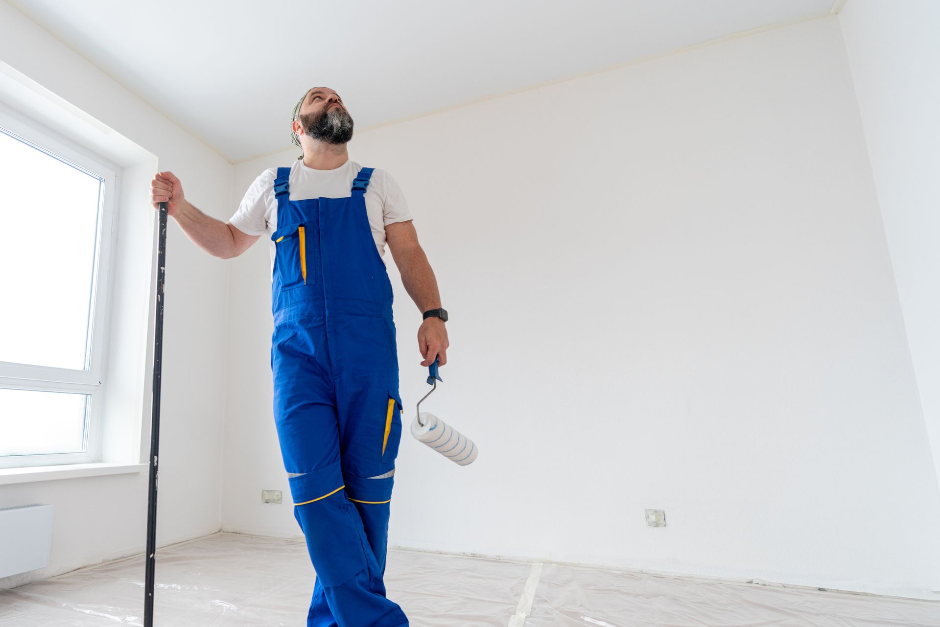 Painter in blue overalls holding a paint roller in a bright, empty room