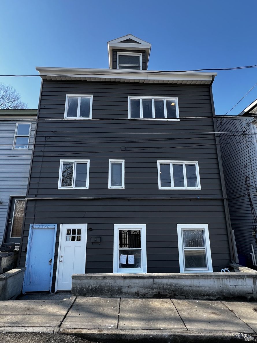 Three-story dark gray house with white-trimmed windows, a blue door, and a small top dormer on a sidewalk.