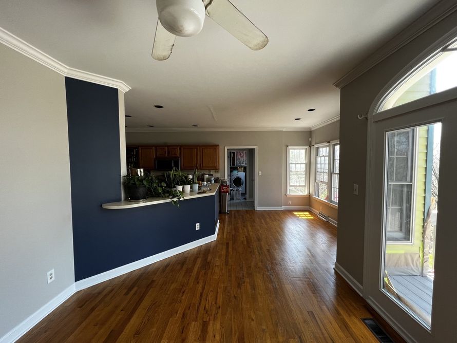 Empty living room with hardwood floors, dark blue accent wall, open kitchen, and large arched window.
