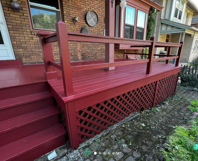 Red painted wooden deck with lattice skirting, stairs, and railing, in front of a brick house.