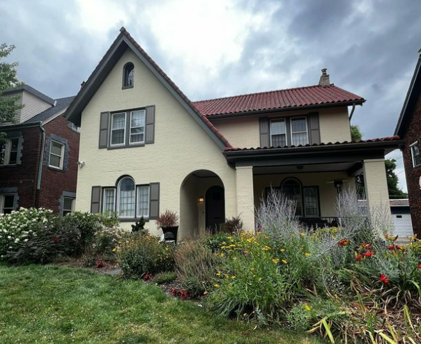 Two-story beige house with red tile roof, front porch, and garden under a cloudy sky.