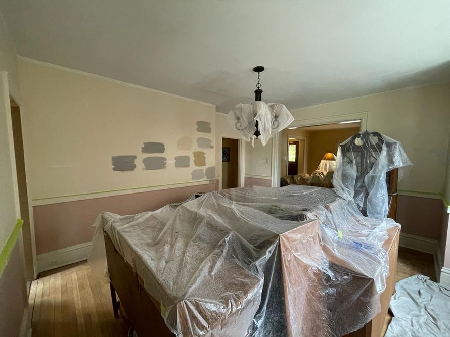 Living room covered in plastic sheeting during painting or renovation, with furniture wrapped and wall color samples visible